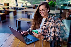 young female in plaid shirt talking into a phone, wearing ear buds and sat in front of a laptop