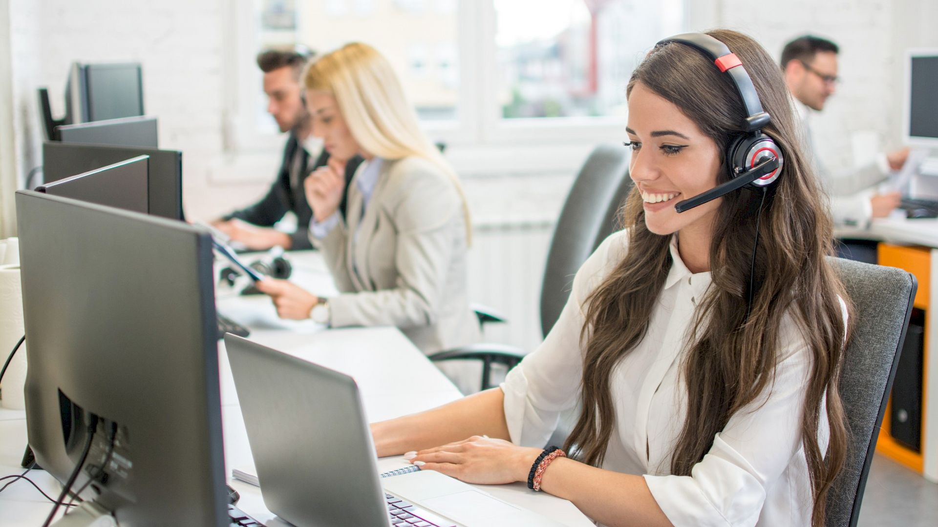 Young female customer service agen wearing a headset an smiling in front of computer screen
