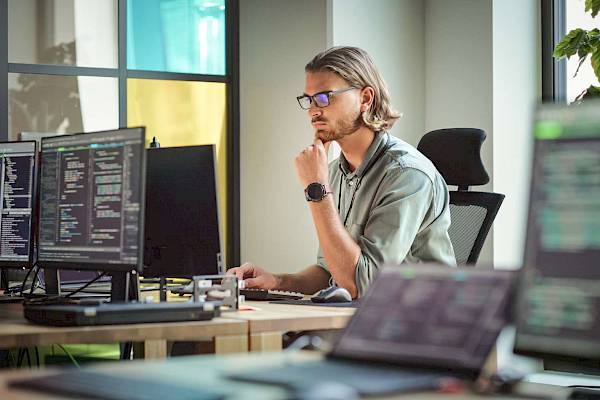 Male computer scientist wearing glasses looking at multiple computer screens with code on